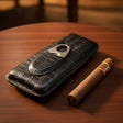 Black crocodile-patterned cigar case with a silver cutter and a cigar on a wooden table