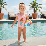 Child in a mermaid swimsuit holding a beach ball by a pool with plants and chairs in the background.