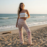 Woman in a white top and patterned pants standing on a beach with ocean waves in the background.