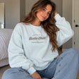 Woman in a light blue crew neck sweatshirt with a vintage script chest print, sitting in a bedroom