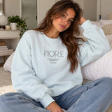Woman in a light blue crew neck sweatshirt with minimalist Fiore lettering, worn with light jeans