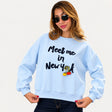 Woman in sunglasses wearing a light blue Meet me in New York sweatshirt against a white background