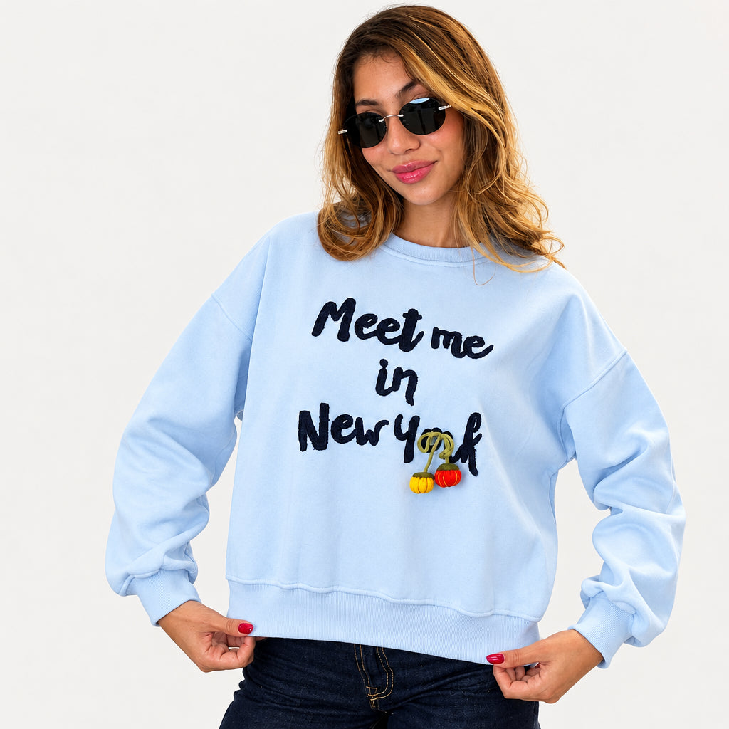 Woman in sunglasses wearing a light blue Meet me in New York sweatshirt against a white background