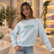 Woman in a light blue sweatshirt with mint sequin lettering, one hand at her jeans pocket