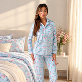 Front view of a light blue rose print button up pyjama shirt and trousers by a white bedside table