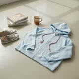 Light blue zip hoodie laid flat on a cream floor with pink tassel drawstrings