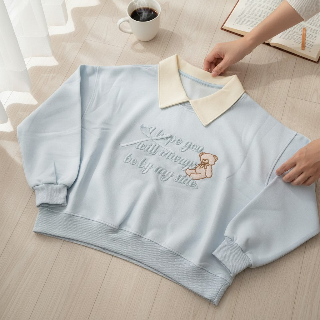 Woman wearing a light blue collared sweatshirt with an embroidered teddy bear and script slogan