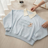 Woman wearing a light blue collared sweatshirt with an embroidered teddy bear and script slogan