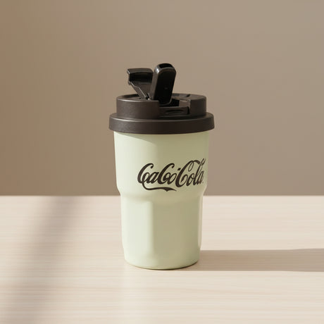 Light green Coca-Cola tumbler with black lid on a white surface, with clothing in the background.