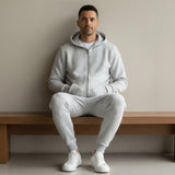 Man wearing a grey tracksuit sitting on a wooden bench against a plain wall
