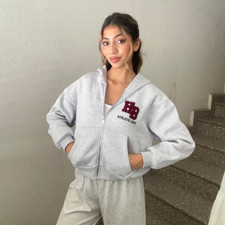 Woman in a light grey cropped zip hoodie with a burgundy letter patch, hands in the front pockets