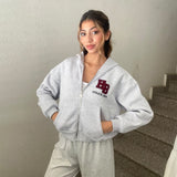 Woman in a light grey cropped zip hoodie with a burgundy letter patch, hands in the front pockets