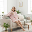 Woman in a pink hooded blanket sitting on a couch holding a mug in a bright living room.