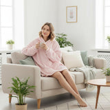 Woman in a pink hooded blanket sitting on a couch holding a mug in a bright living room.