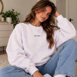 Woman in a lilac crew neck sweatshirt with a dainty Aube de Paris script, hands in her jeans pockets