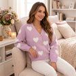 Woman in a lilac V neck cardigan with white hearts and large buttons, worn with white jeans in a cosy room