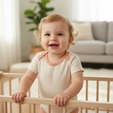Close view of a baby at a crib rail in a cream bodysuit with fine pink stripes and peach neck binding