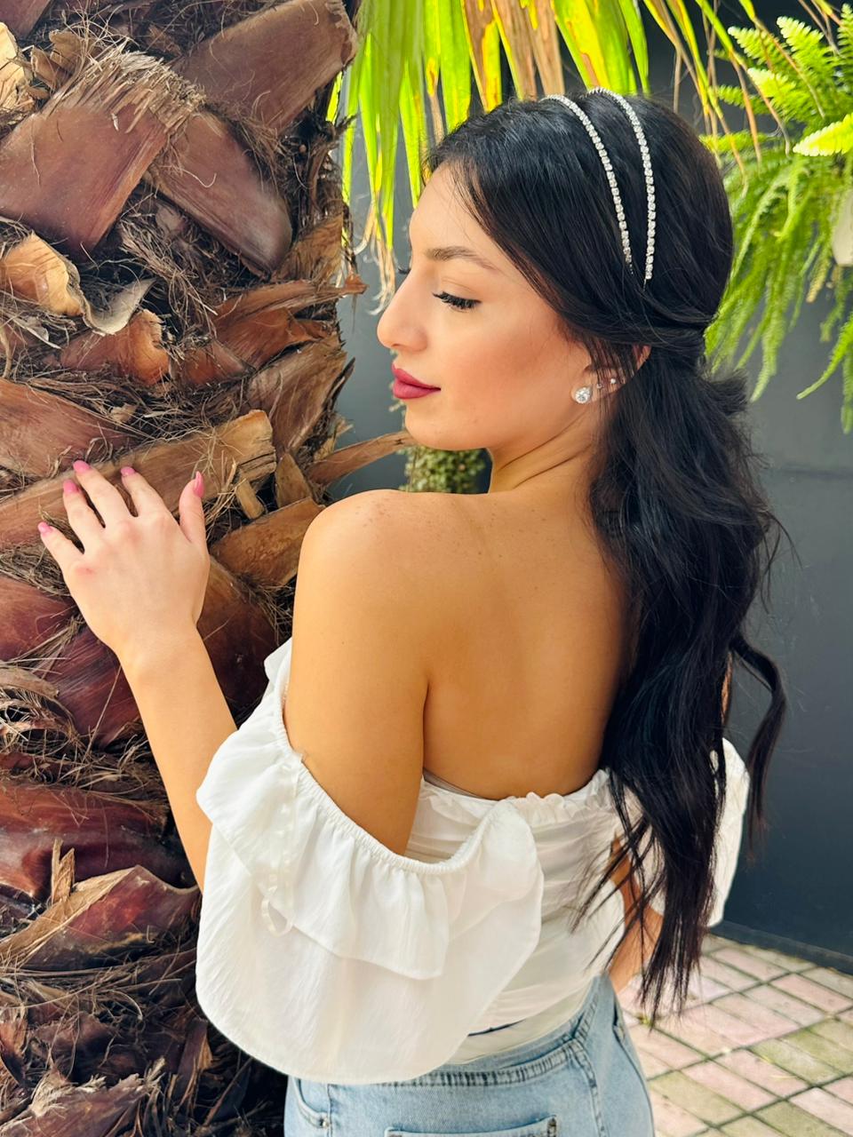 Woman wearing a luxe charm crown accessory in a white off-shoulder top standing next to a palm tree