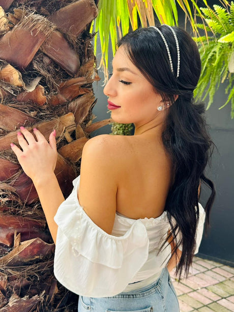Woman wearing a luxe charm crown accessory in a white off-shoulder top standing next to a palm tree