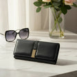 Black wallet with gold accent on a white surface, accompanied by sunglasses and a vase of flowers