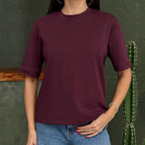Person wearing a maroon red tshirt and blue jeans standing against a green wall with a cactus plant
