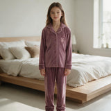 Girl standing barefoot in a mauve velvet pyjama set with pink piping beside a bed