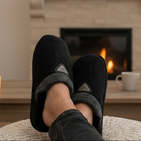 Person wearing black slippers with a fireplace and mug in the background
