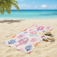 Beach towel with mermaid and jellyfish pattern on a sandy beach with ocean and palm tree in the background.
