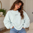 Woman in a mint crew neck sweatshirt with a dainty Aube de Paris script, hands in her jeans pockets