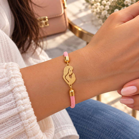 Pink braided cord bracelet with a gold mother and baby silhouette charm, pink bag behind