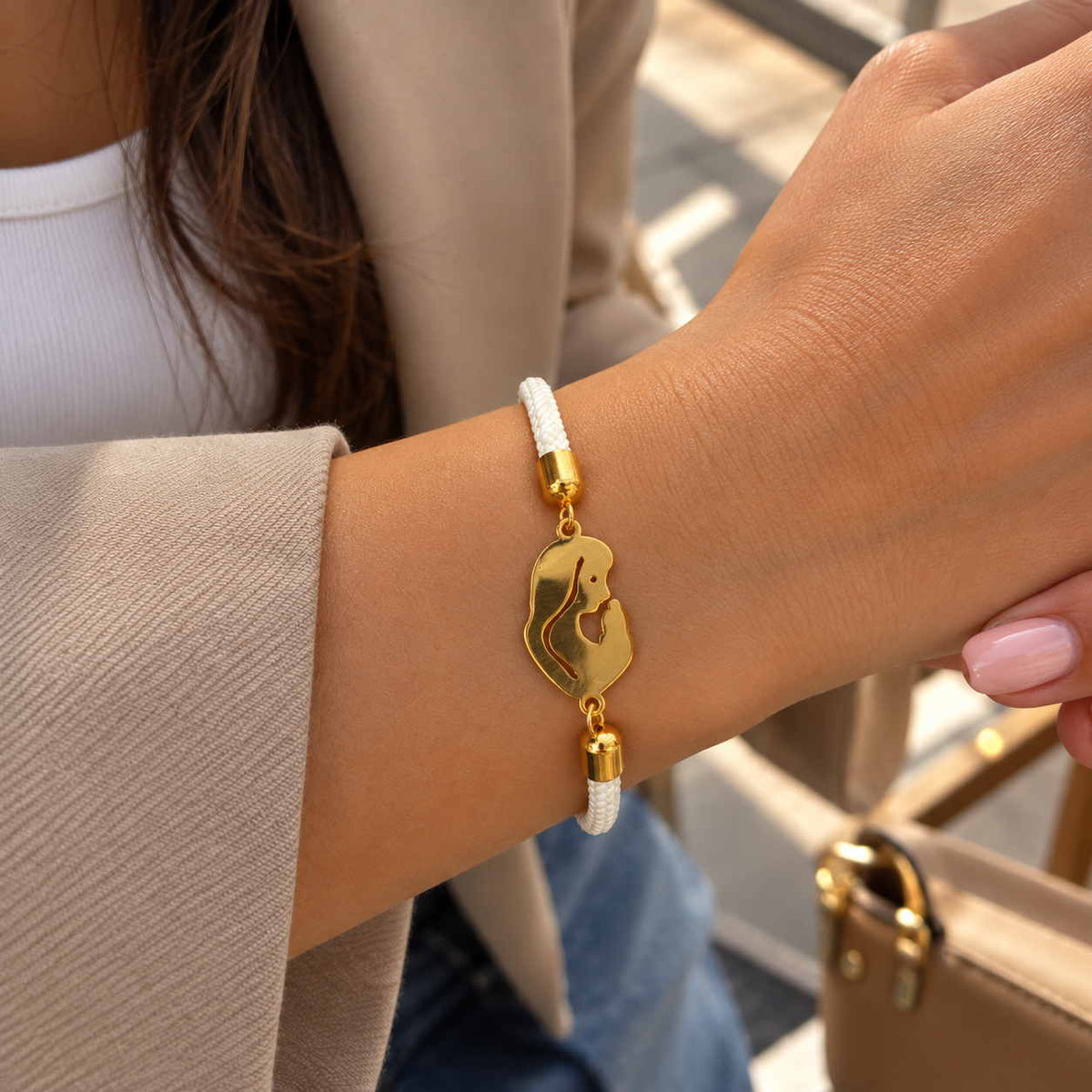Wrist wearing a white braided cord bracelet with a gold mother and child charm, beige blazer sleeve
