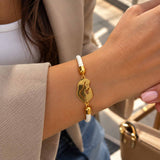 Wrist wearing a white braided cord bracelet with a gold mother and child charm, beige blazer sleeve