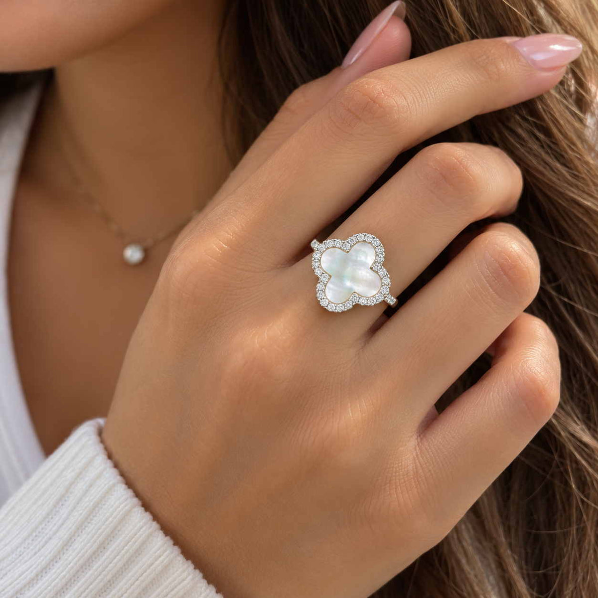 Hand wearing a ring with a large pearly white clover framed by small crystals, fingers in long hair