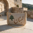 Light linen pouch with Lebanon stitched in bronze Arabic beside a green cedar, on a stone terrace near an old arch
