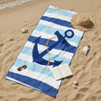 A beach towel laid on sandy shore with a large navy anchor and blue-and-white striped pattern. A straw hat, sunglasses, open book, and small bottle are placed around it as lifestyle props.