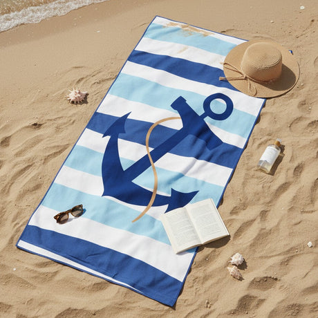 A beach towel laid on sandy shore with a large navy anchor and blue-and-white striped pattern. A straw hat, sunglasses, open book, and small bottle are placed around it as lifestyle props.