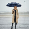 Woman holding a navy blue umbrella in the rain wearing a beige trench coat