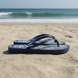 Navy blue flip-flops with white patterns on a sandy beach with ocean waves in the background