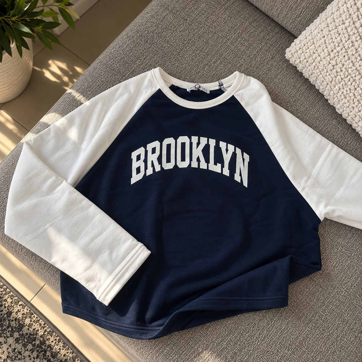 Navy Brooklyn sweatshirt with white raglan sleeves laid flat on a grey sofa