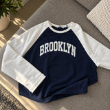 Navy Brooklyn sweatshirt with white raglan sleeves laid flat on a grey sofa