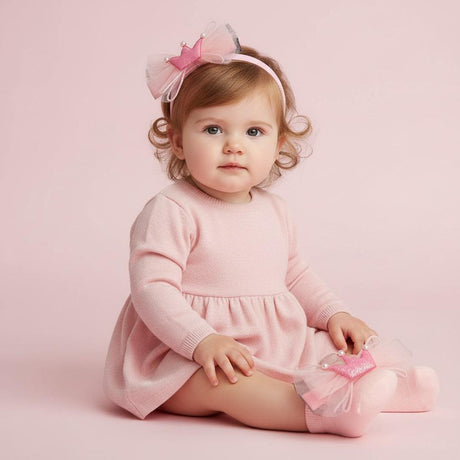 Seated baby in a pink knit dress with a tulle crown headband and soft pink socks with crown appliques