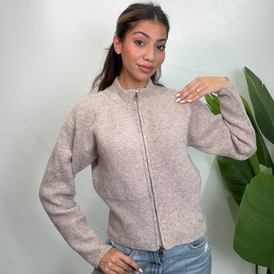 Woman in a heathered oatmeal knit cardigan with one hand on her shoulder by a green plant