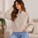 Woman in a heathered oatmeal zip up knit cardigan with a funnel neck sitting on a sofa