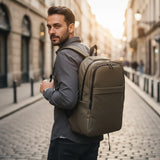 A man wearing a grey long-sleeve shirt and dark jeans carries a large olive backpack on his back while standing on a cobblestone street
