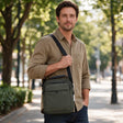 Man in a khaki shirt wearing an olive crossbody bag with a zip front pocket on a tree lined street
