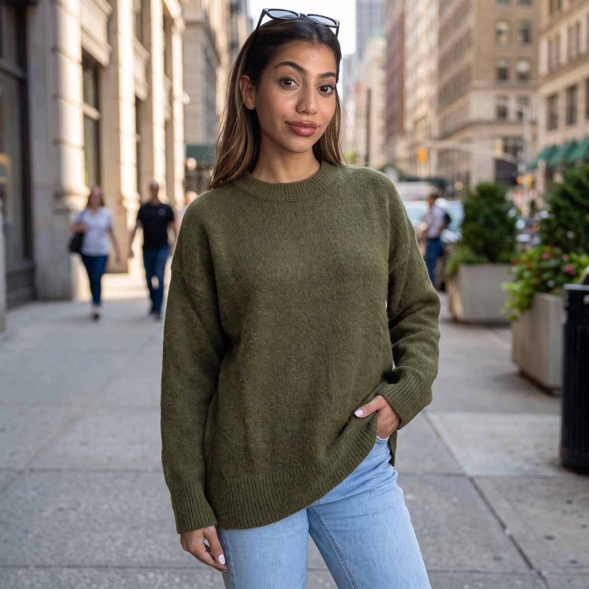 Woman in an olive green brushed wool crew neck sweater and light jeans on a city pavement, hand in her pocket