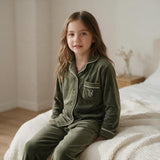 Girl sitting on a bed in olive green velvet pyjamas showing the embroidered chest pocket