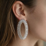 Silver open oval earring lined with clear baguette crystals worn on the ear