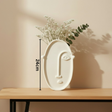 White face-shaped vase with plants on a wooden table against a beige wall