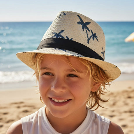 Child wearing a beige woven straw fedora with navy palm trees and a black band on a shore